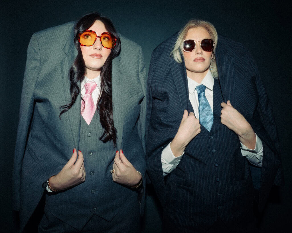 Two professional women in suits with oversized jackets, wearing sunglasses, posing confidently.